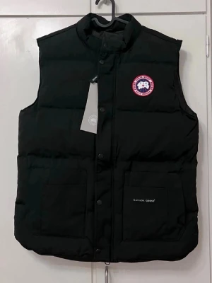Svart dunväst från Canada Goose - Snygg svart dunväst från Canada Goose med hög krage, tryckknappar och två stora framfickor. Klassisk logopatch på bröstet och diskret broderad logga på fickan. Perfekt för lager-på-lager och håller dig varm under kyliga dagar. S till L storlekar finns