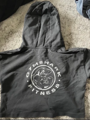 Svart cropped hoodie från Gymshark - Säljer en svart cropped hoodie från Gymshark med stort tryck på ryggen där det står 'Gymshark Fitness' och en haj-logga. Hoodien har huva med dragsko och korta ärmar. Aldrig använd. 