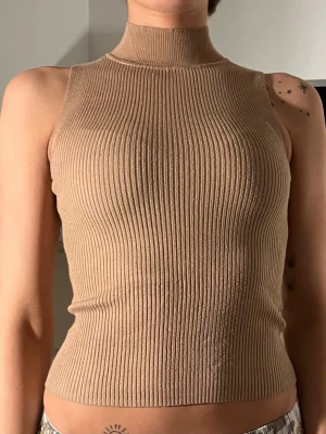 Beige ribbad ärmlös topp - Snygg beige ribbad topp med hög hals och ärmlös design. Toppen är figurnära och har en stretchig, stickad känsla som gör den bekväm och trendig. Perfekt att styla med jeans eller kjol för en clean look.