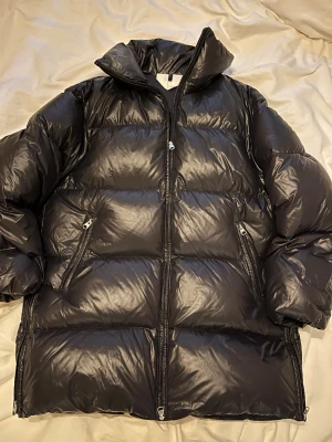 Svart pufferjacka med hög krage ARKET - Säljer en svart pufferjacka med hög krage och två dragkedjeförsedda fickor framtill. Jackan har en glansig finish och breda, vadderade paneler som ger en riktigt puffig look. Perfekt för kalla vinterdagar och riktigt snygg till streetwear-stilen. Använd men i fint skick. Har sminkfläckar som jag inte försökt ta bort.