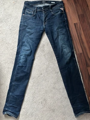 Mörkblå Replay Anbass slim fit jeans - Säljer ett par mörkblå jeans från Replay, modell Anbass. Jeansen har klassisk femficksdesign, smal passform och snygga slitningar framtill. Tillverkade i stretchigt denimtyg för extra komfort. Perfekta för dig som gillar en modern och stilren look.