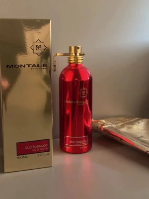Montale Oud Tobacco Eau de Parfum - Montale Oud Tobacco Eau de Parfum i en metallicröd cylinderformad flaska med gulddetaljer. Flaskan rymmer 100 ml och kommer med en matchande guldig förpackning och påse. Doften är exklusiv och flaskan har en modern, lyxig design. Säljer den då jag inte använder ofta, den är iprincip helfull, kontakta gärna vid intresse🤩🙏🏼
