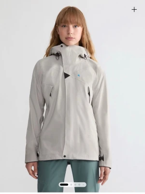 Ljusgrå vindjacka från Klättermusen - Snygg ljusgrå vindjacka från Klättermusen med justerbar huva och smarta detaljer som snedställd dragkedja och ventilationsöppning. Jackan har två fickor framtill och diskret logga på bröstet. Perfekt för dig som gillar outdoor och stilren design. Helt ny prislappen kvar 