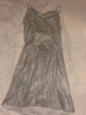 Glittrig silver kortklänning med snörning - Supersnygg glittrig silverfärgad kortklänning med tunna axelband och öppen rygg med snörningsdetalj. Klänningen har en lätt utsvängd form och är perfekt för dig som vill sticka ut. Materialet är glittrigt och ger en festlig känsla.