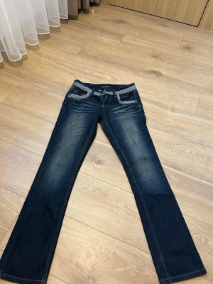 Mörkblå bootcut jeans från M.A.P - Säljer ett par mörkblå bootcut jeans från M.A.P med snygga slitningar och kontrastsömmar. Jeansen har dekorativa bakfickor med broderi och detaljer i grått vid fickor och linning. Lågmidjade 