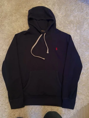 Marinblå hoodie från Polo Ralph Lauren - Snygg marinblå hoodie från Polo Ralph Lauren med klassisk röd logga broderad på bröstet. Hoodien har vit dragsko, stor känguruficka och ribbade muddar. Tillverkad i mjuk bomull för maximal komfort och stil.
