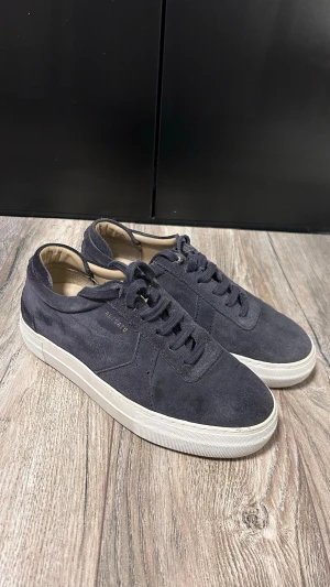 Mörkblå mockasneakers från Arigato - Stilrena mörkblå sneakers i mocka från Arigato med klassisk låg profil och vita sulor. Skorna har matchande mörkblå skosnören och diskret logga på sidan. Perfekta för dig som gillar en clean och enkel look.