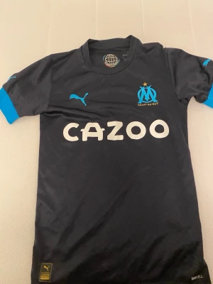 Marseille fotbollströja Puma svart - Snygg svart fotbollströja från Puma med Olympique de Marseille-logga och sponsortryck 'Cazoo' på bröstet. Tröjan har ljusblå detaljer på ärmarna och axlarna samt Drycell-material för bra ventilation. Perfekt för träning eller match!