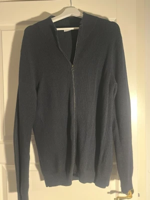 Mörkblå ribbad ziptröja - Mörkblå ribbstickad tröja med dragkedja framtill och lång ärm. Tröjan har en enkel, stilren design utan synliga loggor eller detaljer och är tillverkad i ett mjukt stickat material. Perfekt för lager på lager och chill vardagsstil.