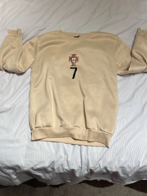 Beige Ronaldo tröja Portugal nr 7 - Beige sweatshirt med Portugals landslagsemblem och nummer 7 på framsidan. På baksidan står det 'RONALDO' och en stor svart 7:a. Tröjan har rund hals och ribbade muddar vid ärmslut och nederkant. Perfekt för dig som gillar fotboll och Cristiano Ronaldo.