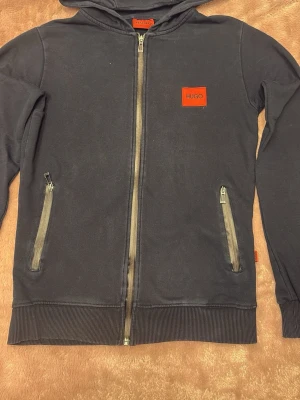 Mörkblå hoodie från HUGO - Snygg mörkblå hoodie från HUGO med dragkedja framtill och två fickor med dragkedja. Hoodien har en röd HUGO-logga på bröstet och matchande röd etikett i sidan. Tillverkad i mjukt bomullsmaterial och har ribbade muddar vid ärmslut och nederkant. Färgen har släppt lite på metallringarna där snöret går och smöret saknas. Annars i mycket bra skick. Se bilderna. 