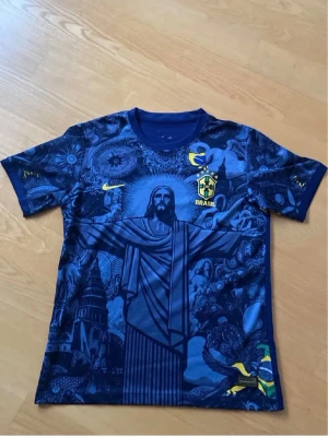 Blå Brasilien t-shirt Nike - Unik blå t-shirt från Nike med Brasilien-tema och detaljerat tryck av Kristusstatyn och brasilianska symboler. T-shirten har korta ärmar, rund hals och Brasil-logga på bröstet. Perfekt för dig som vill sticka ut med en sportig och cool look.