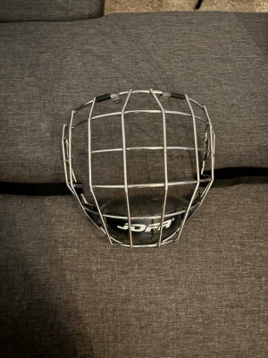 Jofa hockeygaller i metall 480M - Säljer ett klassiskt hockeygaller från Jofa i silverfärgad metall med svart plastdetalj och justerbart band. Gallret har en robust konstruktion och passar till hjälm för extra ansiktsskydd under hockey eller liknande sporter.