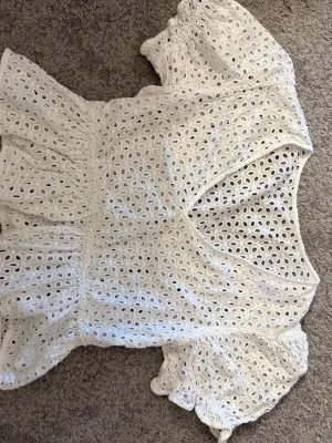 Vit broderad blus med puffärm - Söt vit blus i broderad bomull med v-ringning och korta puffärmar. Blusen har elastisk midja och är dekorerad med hålmönster över hela plagget. Perfekt för en somrig och fräsch look.