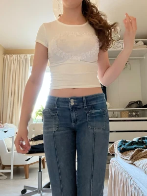 Vit croppad t-shirt med hjärtdetalj - Söt vit croppad t-shirt med korta ärmar och ett hjärta i volang framtill. Tillverkad i mjuk bomull och perfekt för en trendig och avslappnad stil. Passar dig som gillar gulliga detaljer och enkel design.