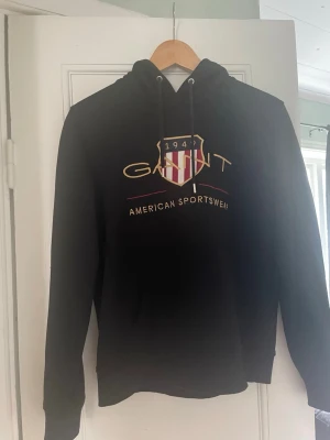 Svart hoodie från Gant med tryck - Snygg svart hoodie från Gant med stort tryck på bröstet i guld, vitt och rött. Klassisk huva med dragsko och ribbade muddar. På framsidan står det 'GANT 1949 American Sportswear' och ett sköldmärke. Perfekt för en sportig och avslappnad stil.