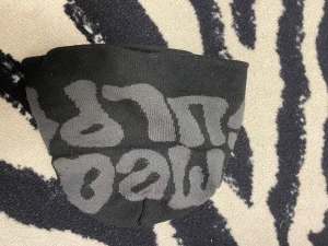 Svart mössa med grå text - Säljer en svart mössa med stor grå text i ett grafiskt mönster runt om. Mössan är stickad och har en avslappnad passform, perfekt för kalla dagar. Materialet känns mjukt och stretchigt, vilket gör den bekväm att bära hela dagen.