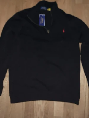 Svart halvzip tröja från Polo Ralph Lauren - Klassisk svart halvzip tröja från Polo Ralph Lauren med den ikoniska röda broderade loggan på bröstet. Tröjan har lång ärm, ribbade muddar och hög krage med dragkedja. Perfekt för en stilren och avslappnad look.