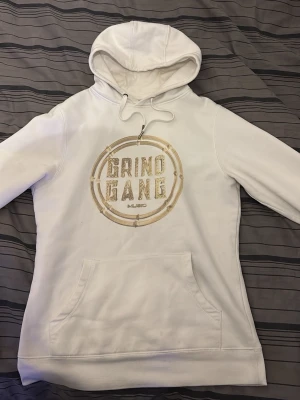 Vit hoodie Grind Gang Music - Snygg vit hoodie med guldigt Grind Gang Music-tryck på bröstet. Klassisk känguruficka framtill och justerbar huva med dragsko. Perfekt för dig som gillar streetwear och vill sticka ut med en unik design. Tröjan har två små hål vid fickan men syns inte när man har på si och enkelt att fixa om man vil göra de, BYTEN KAN OCKSÅ VARA INTRESSANT!!
