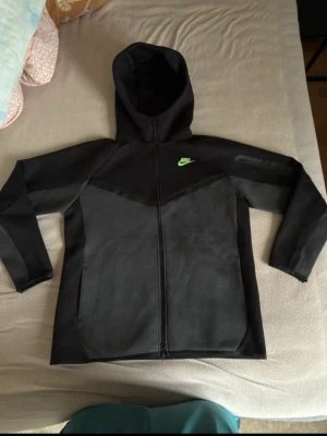 Nike tech fleece - Snygg svart ziptröja från Nike med hög krage och neongrönt Nike-märke på bröstet. Tröjan har dragkedja framtill, en ficka med dragkedja på ärmen och är gjord i ett mjukt, stretchigt material. Perfekt för en sportig och avslappnad stil.
