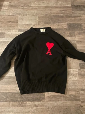 Ami Paris tröja - Svart stickad tröja från Ami Paris med röd broderad hjärta och bokstaven A på bröstet. Tröjan har rund halsringning, ribbstickade muddar och är tillverkad i mjukt material. Perfekt för dig som gillar stilrena detaljer och ikoniska loggor.