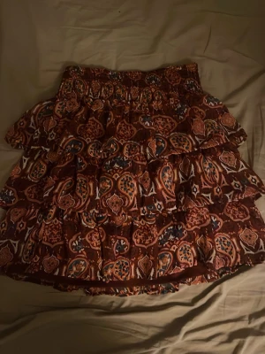 Brun mönstrad volangkjol från Lindex - Kort volangkjol från Lindex i brunt med paisleyinspirerat mönster i orange, blått och vitt. Kjolen har bred resår i midjan och flera lager volanger som ger en flowy känsla. Perfekt för dig som gillar boho-vibbar och färgstarka plagg.