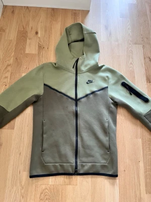 Nike Tech Fleece hoodie i olivgrönt - Nike Tech Fleece hoodie i två nyanser av olivgrönt med hel dragkedja och svart Nike-logga på bröstet. Hoodien har fickor framtill, en extra ficka med dragkedja på ärmen och svarta detaljer längs dragkedja och muddar. Tillverkad i mjukt och bekvämt material.