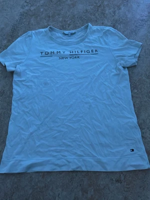 Vit Tommy Hilfiger t-shirt - Snygg vit t-shirt från Tommy Hilfiger med tryck 'Tommy Hilfiger New York' på bröstet och liten logga nere vid fållen. Klassisk rund hals och korta ärmar. Perfekt till jeans eller shorts för en clean och enkel look. Det är en liten fläck nertill men den syns knappt