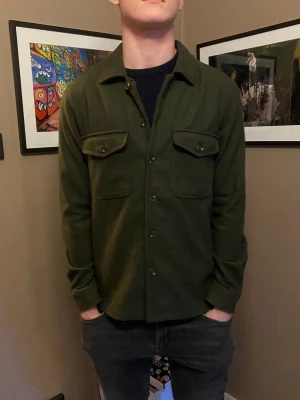 Mörkgrön overshirt med fickor - Stilren mörkgrön overshirt med knappar framtill och två bröstfickor med lock. Skön passform och klassisk krage, perfekt att bära över en t-shirt. Materialet ser ut att vara mjukt och lite tjockare, vilket ger en avslappnad look.