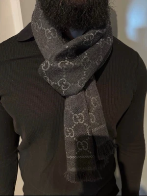 Mörkgrå halsduk från Gucci - Snygg mörkgrå halsduk från Gucci med det klassiska GG-mönstret i ljusgrått. Halsduken har fransade kanter och är tillverkad i ett mjukt material som känns lyxigt. Perfekt accessoar för att lyfta din outfit.