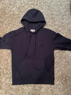 Mörkblå hoodie - Snygg mörkblå hoodie med broderad CD-logga på bröstet. Klassisk modell med huva och dragsnören samt en stor magficka framtill. Tillverkad i mjukt bomullsmaterial som känns skönt mot huden. Perfekt för en avslappnad och trendig look.