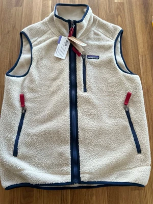 Beige fleeceväst från Patagonia - Snygg beige fleeceväst från Patagonia med blå detaljer och röda dragkedjor. Västen har hög krage, tre fickor med dragkedja och är gjord i mjukt, fluffigt material. Perfekt för lager-på-lager och outdoor-stil.