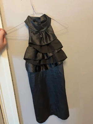 Festtopp med volanger - Snygg svart halterneck topp i satin med flera lager volanger framtill. Toppen är ärmlös och har en hög krage som knäpps i nacken. Perfekt för dig som vill sticka ut med en elegant och trendig look. Hade endast bilder på från en gammal TikTok video så man kan ksk inte ser så bra…