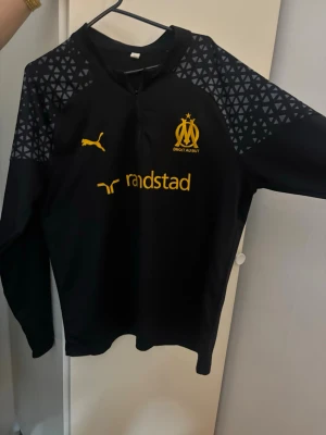 OM träningströja svart Puma M - Det står att det är storlek XL men det är storlek M Snygg svart långärmad träningströja från Puma för Olympique de Marseille. Tröjan har gula loggor och tryck, samt grått mönster på axlarna. Perfekt för fotbollsträning eller chill. Materialet är troligtvis polyester för bästa komfort.