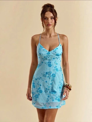 Blå blommig kort klänning med snörning - Supersöt ljusblå kort klänning med blommigt, paljett & glitter mönster och tunna axelband som korsas och knyts i ryggen. Klänningen har v-ringning och är ärmlös, perfekt för varma dagar. Materialet känns lätt och luftigt, vilket gör den idealisk för sommaren.