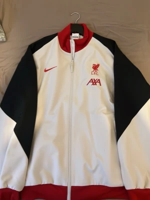 Liverpool Nike vit träningsjacka - Snygg vit träningsjacka från Nike med Liverpool FC-logga och AXA sponsortryck på bröstet. Jackan har svarta ärmar, röda detaljer vid krage och nederkant samt hel dragkedja framtill. Perfekt för dig som älskar fotboll och vill ha en sportig look.