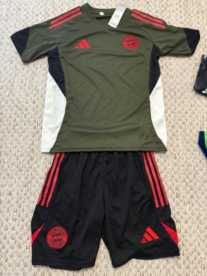 FC Bayern München träningsset Adidas - Snyggt träningsset från Adidas med FC Bayern München-logga. Tröjan är olivgrön med röda och svarta detaljer samt vita sidopaneler. Shortsen är svarta med röda ränder och logga. Perfekt för fotbollsträning eller gymmet.