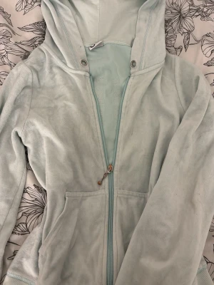 Ljusblå hoodie med paljetter - Mysig 2000-talet ljusblå hoodie i mjukt velourmaterial med dragkedja framtill och fickor. På ryggen står det 'TIME OFF' i stora bokstäver dekorerade med blå och gröna paljetter. Perfekt för chill dagar och avslappnad stil. Huva med tryckknappar vid halsen.