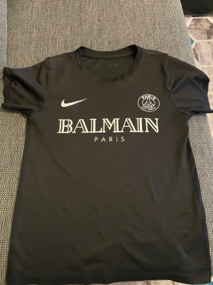 Svart PSG x Balmain t-shirt Nike - Snygg svart t-shirt från Nike i samarbete med PSG och Balmain. T-shirten har korta ärmar, rund halsringning och tryck framtill med Balmain Paris och PSG-logga. Tillverkad i mjuk polyester som känns skön mot huden.