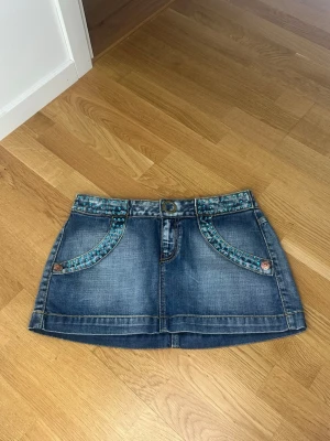 Blå jeanskjol med strass - Säljer en blå jeanskjol med kort längd och snygga strass vid fickorna och linningen. Kjolen har två framfickor, två bakfickor och knappstängning. Perfekt för dig som gillar denim med en twist!