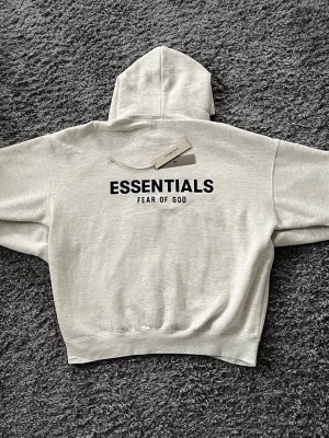 Grå Essentials Fear of God hoodie - Snygg ljusgrå hoodie från Essentials Fear of God med stor svart logga på ryggen. Klassisk oversized passform med huva och ribbade muddar. Perfekt för dig som gillar streetwear och vill ha en stilren look. Materialet känns mjukt och bekvämt.