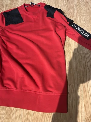 Röd sweatshirt från Moncler - Röd sweatshirt från Moncler med svarta detaljer på axlarna och ett stort Moncler-tryck på ena ärmen. Tröjan har rund halsringning och är tillverkad i ett mjukt bomullsmaterial. Perfekt för en streetwear-look.