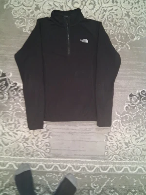 Svart fleece från The North Face - Svart fleece med halv dragkedja från The North Face. Tröjan har hög krage och logga broderad på bröstet. Perfekt för lager-på-lager och enkel att matcha med andra plagg. Långärmad och mjuk i materialet.