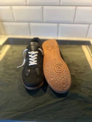 Svarta sneakers med vita snören, gata - Stilrena svarta sneakers med vita skosnören och brun sula. Ovandelen är i en mix av mocka och textil, vilket ger en klassisk look. Perfekta för dig som gillar minimalistisk och tidlös stil.