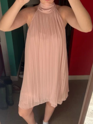 Ljusrosa plisserad halterneckklänning - Säljer en ljusrosa kortklänning med plisserat tyg och halterneck med knyt i nacken. Klänningen är ärmlös och har en lös passform som ger ett flowy intryck. Perfekt för sommarens alla tillfällen! Jag är 165 som referens