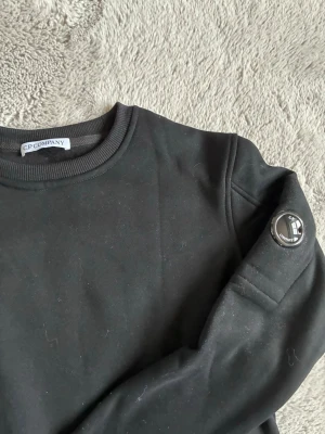 Svart sweatshirt från C.P. Company - Säljer en svart sweatshirt från C.P. Company med rund halsringning och ribbade muddar. Tröjan har den ikoniska linsen på ärmen och är tillverkad i mjukt bomullsmaterial. Perfekt för en clean och stilren look.