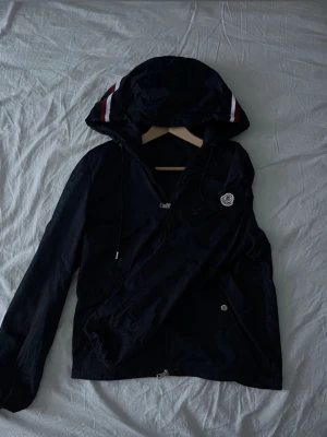 Mörk blått Moncler vindjacka med huva - Säljer en svart vindjacka från Moncler med huva och vita/röda detaljer på huvan. Jackan har dragkedja framtill, två fickor med knappar och Moncler-logga på ärmen. Perfekt för blåsiga dagar och snygg att matcha med jeans. Pris kan bli diskuterat