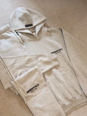 Grå mjukisbyxor Essentials Fear of God - Snygga grå mjukisbyxor från Essentials Fear of God med svart logotyp på benet. Byxorna har resår i midjan och vid benslut, samt dragsnöre för justering. Perfekt för en avslappnad och trendig look. Materialet är mjukt och bekvämt, idealiskt för chill dagar.
