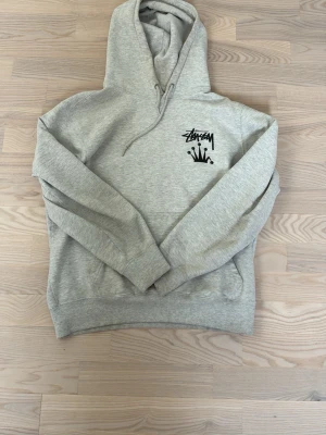  Stüssy hoodie  - Säljer en grå hoodie från Stüssy med svart logga och krona tryckt på bröstet och ryggen. Hoodien har huva med dragsko och ribbade muddar. Tillverkad i mjukt bomullsmaterial, perfekt för en avslappnad streetwear-look.