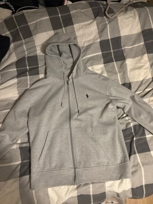 Ljusgrå hoodie från Polo Ralph Lauren - Snygg ljusgrå hoodie med dragkedja från Polo Ralph Lauren. Klassisk design med huva, dragsnören och kängurufickor. Liten broderad logga på bröstet. Perfekt för en chill och stilren look. Den är aldrig använd i princip.